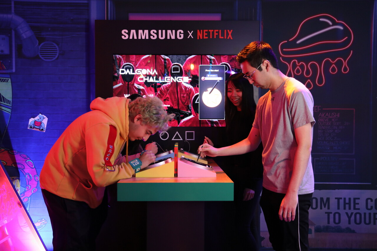 삼성전자는 넷플릭스가 미국 로스앤젤레스에 개장한 체험존인 '오징어게임: 더 트라이얼'의 공식 파트너사로 참여했다. 참가자들이 갤럭시 S23 울트라의 S펜으로 디지털 달고나 게임을 즐기고 있다. 